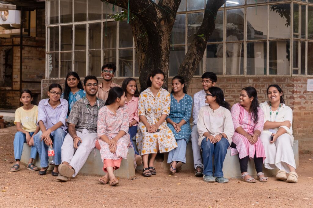 A group of students sitting and laughing together on a low wall under a tree